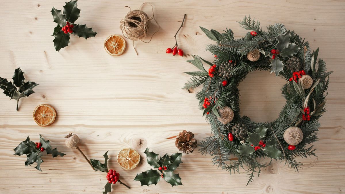 On the table are materials to get ready for Christmas decorations, including leaves, dried lemon slices, red berries, and twine, along with a completed Christmas wreath.