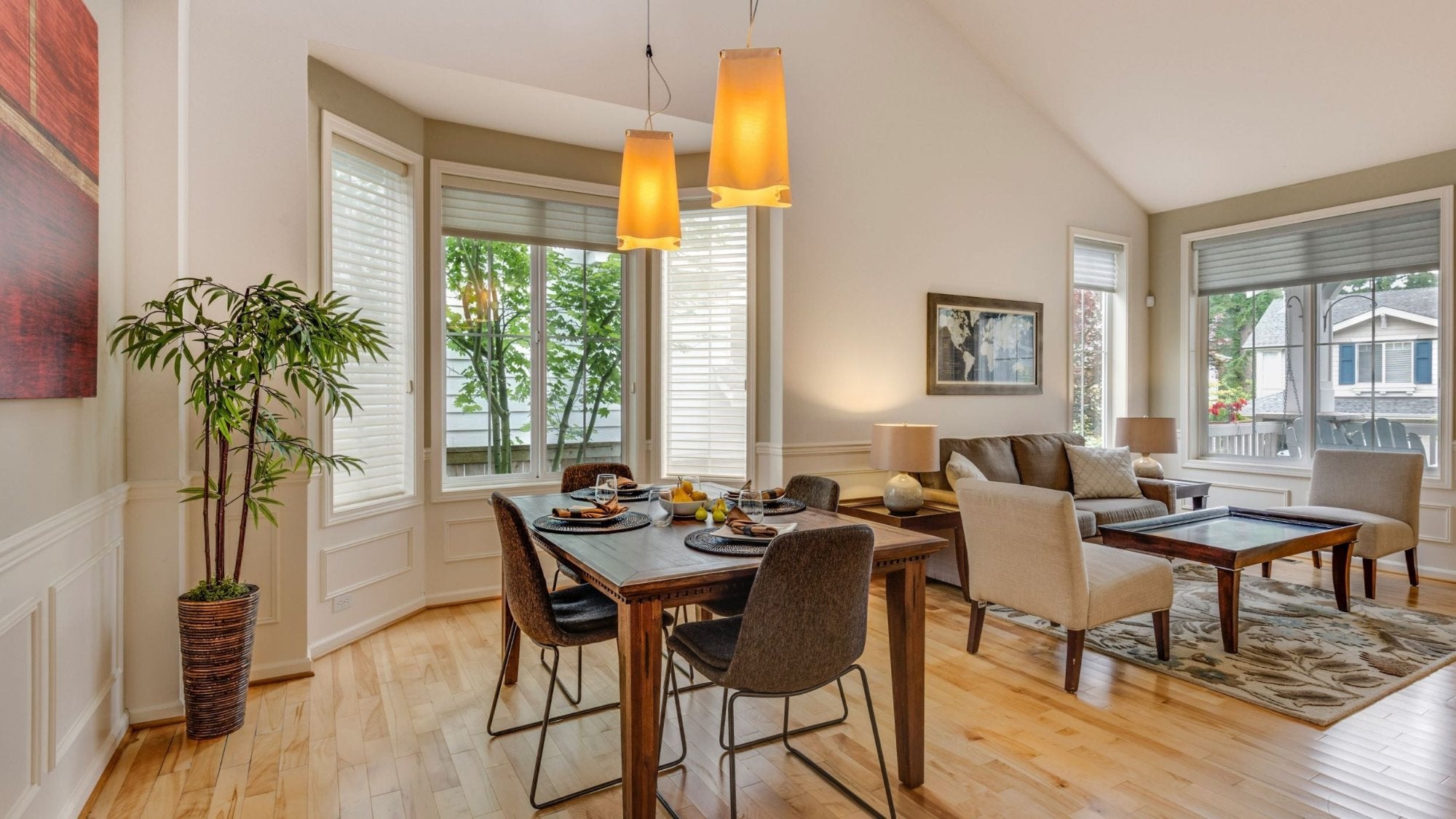 A perfectly balanced living and dining area, featuring window blinds that allow a view of the greenery outside. The dining table is set with a fruit bowl and dinner plates, creating a calm and lively atmosphere in the room.