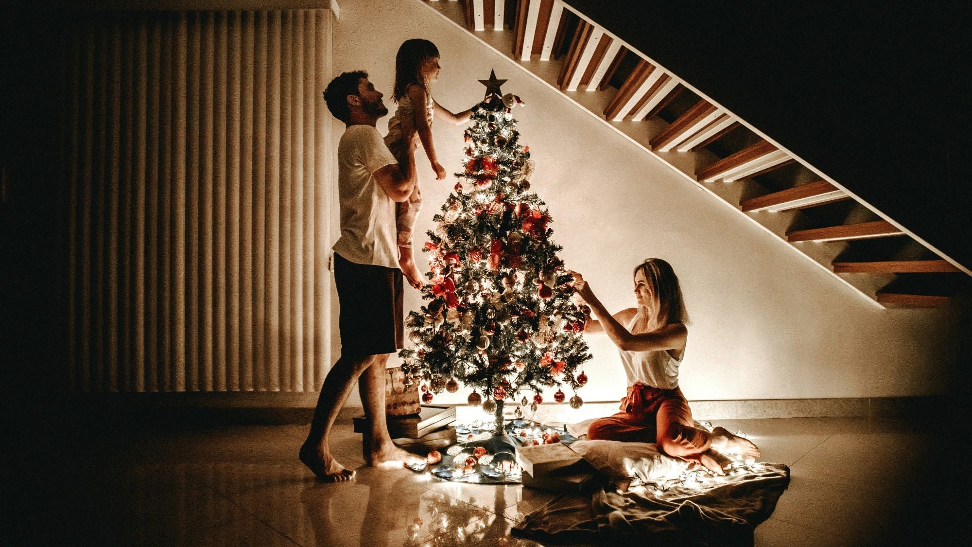 On the ground floor of the house, a family of three is joyfully decorating the Christmas tree. The father is holding his little daughter and placing a star on top of the tree, while the mother is adjusting the lights on the branches.