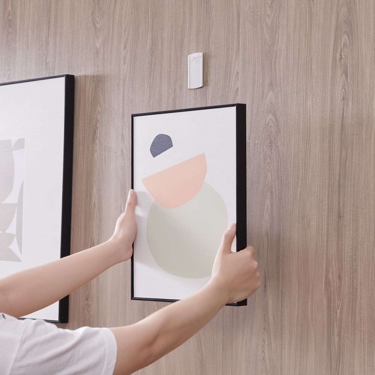 A man holds a picture frame with both hands, preparing to hang it on the wall where a large universal picture hanger is already installed.