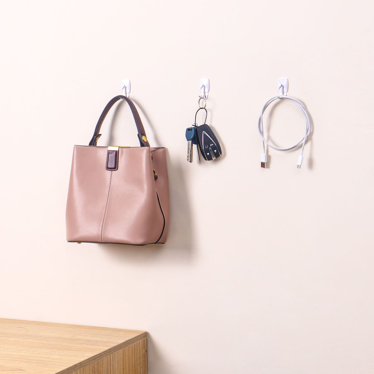 The white hooks are attached to the wall, each holding a pink handbag, a set of keys, and a charging cable.